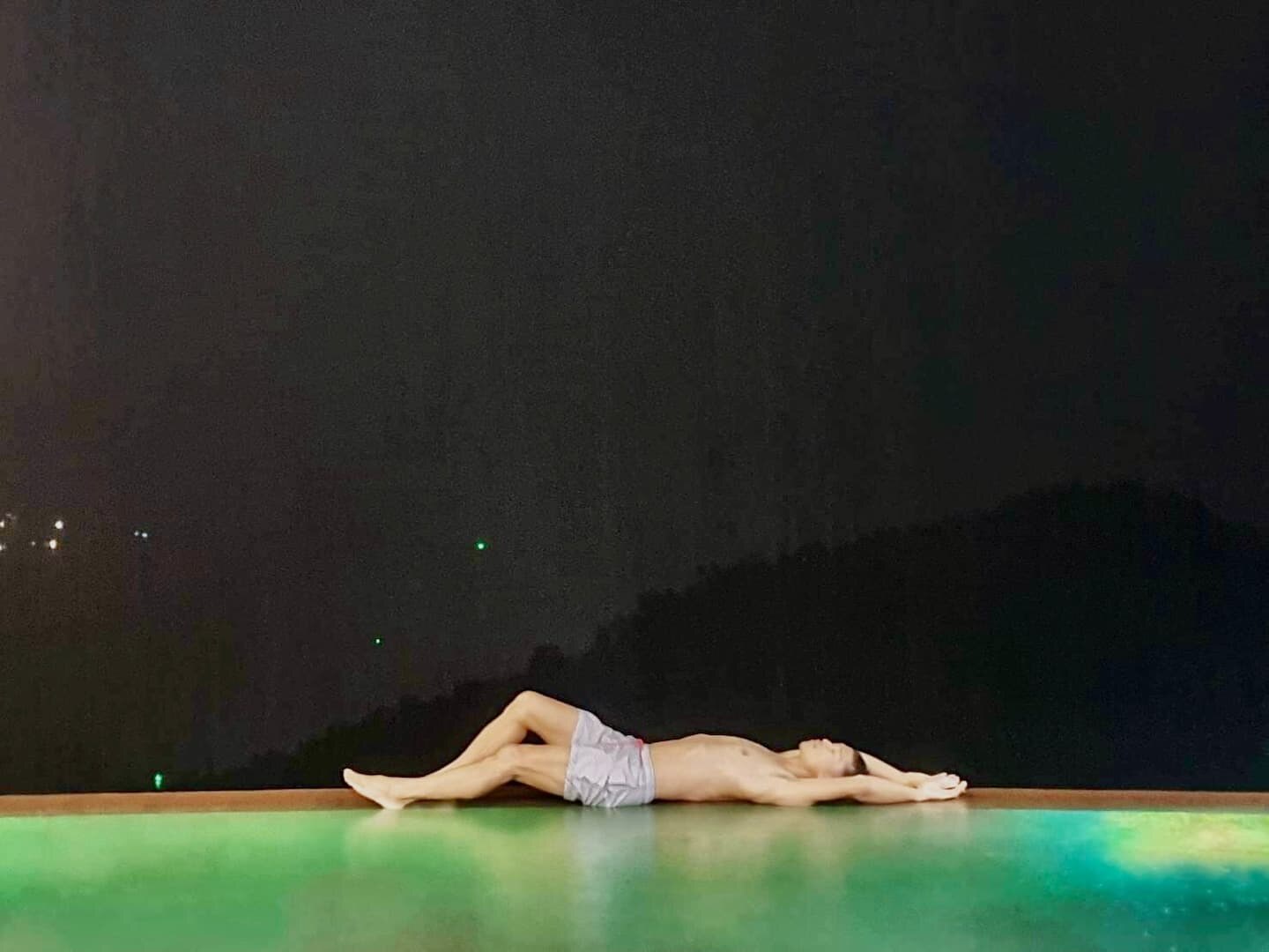 A breathtaking picture of a male and his physique. Lying at the edge of a pool. Stretching his arms above his head, legs bent just slightly. Arching his back just a little bit, so his chest emphasises his bright silhouette against the night behind him.